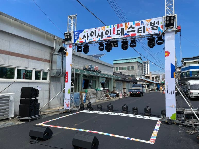 축제 무대·게이트 — 무대 앞 바닥 그래픽과 게이트 배너로 행사 아이덴티티를 입구에서부터 보여줍니다.